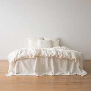 May include: A white linen bed with a white duvet cover and three white pillows. The bed is on a light wood floor.