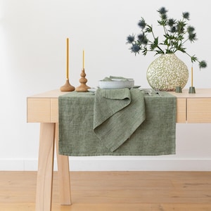 Puede incluir: Un camino de mesa de lino verde con una servilleta doblada sobre una mesa de madera. Hay dos candelabros de madera con velas amarillas y un jarrón con flores secas sobre la mesa.