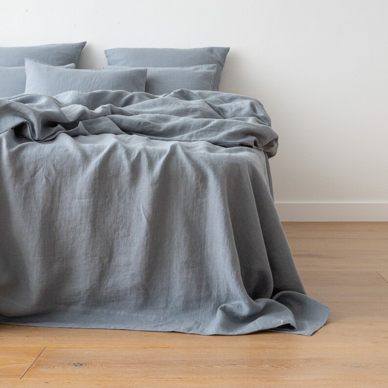 May include: A blue linen bed sheet with a folded linen duvet cover on top. The bed sheet is draped over the edge of the bed and the duvet cover is folded neatly on top.