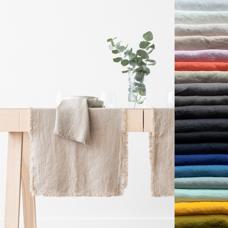 May include: A beige linen table runner with frayed edges, placed on a wooden table with a matching linen napkin. A vase with green leaves sits on the table. The runner is available in a variety of colors.
