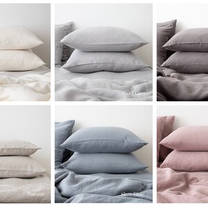 May include: Six linen pillowcases in different colors: natural, cool grey, graphite, dusty rose, slate blue, and natural. The pillowcases are stacked on top of each other on a bed.