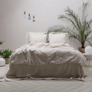 May include: A white bed with a light brown and white striped linen duvet cover and matching pillowcases. The bed is made with a white bed frame and has a white headboard. The bed is in a room with a white wall and a potted plant in the background.