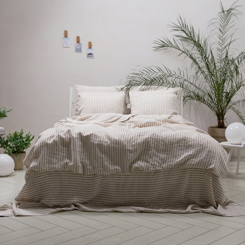 May include: A white bed with a light brown and white striped linen duvet cover and matching pillowcases. The bed is made with a white bed frame and has a white headboard. The bed is in a room with a white wall and a potted plant in the background.