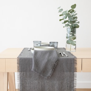May include: A gray linen table runner with frayed edges, set on a wooden table with a gray ceramic bowl, two clear glass cups, and a vase with green eucalyptus branches.