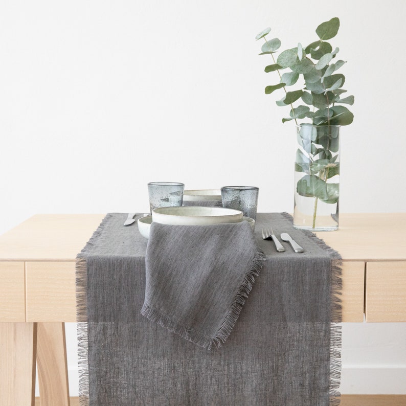 May include: A gray linen table runner with frayed edges, set on a wooden table with a gray ceramic bowl, two clear glass cups, and a vase with green eucalyptus branches.