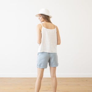 May include: A woman wearing a white linen tank top and light blue linen shorts. She is wearing a white fedora hat and is standing in front of a white wall.