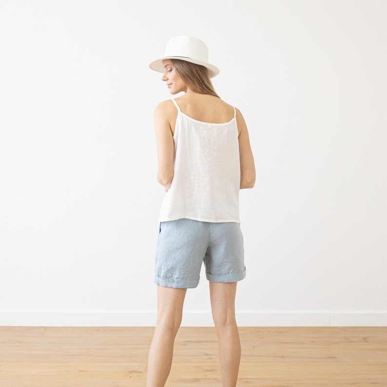 May include: A woman wearing a white linen tank top and light blue linen shorts. She is wearing a white fedora hat and is standing in front of a white wall.