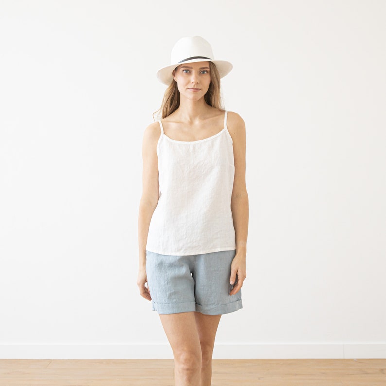 May include: A woman wearing a white linen tank top and light blue linen shorts. She is wearing a white fedora with a black band.