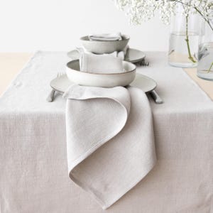 Pode incluir: Uma toalha de mesa de linho cinza claro com guardanapos, tigelas e pratos a condizer. A mesa inclui um guardanapo dobrado, uma tigela e um prato. O fundo inclui um vaso com flores brancas.