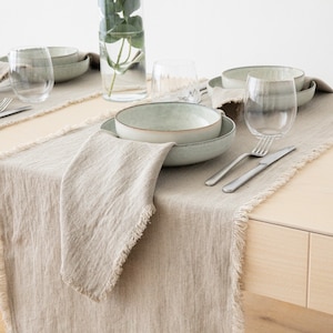 May include: A light brown linen table runner with frayed edges, set on a wooden table. The runner is decorated with a light green ceramic bowl, silverware, and two clear glass wine glasses.