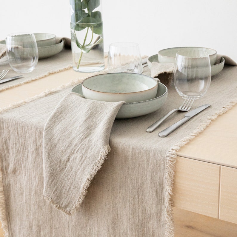 May include: A light brown linen table runner with frayed edges, set on a wooden table. The runner is decorated with a light green ceramic bowl, silverware, and two clear glass wine glasses.
