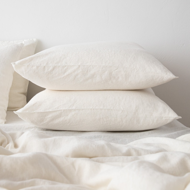 May include: Two white linen pillowcases stacked on top of each other on a white linen bed sheet.