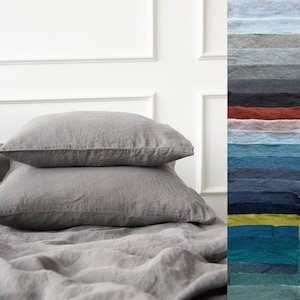 May include: Two gray linen pillowcases stacked on a gray linen bedspread. A swatch card with various colors of linen fabric is shown on the right side of the image.