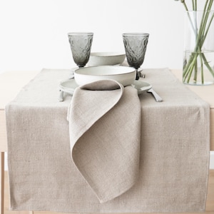 May include: A beige linen table runner with a matching folded napkin, set with white bowls, grey glasses, and cutlery. The table setting is simple and elegant, suitable for a dining room or kitchen.