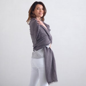 May include: A woman wearing white pants and a gray linen scarf with frayed edges. The scarf is wrapped around her neck and shoulders.