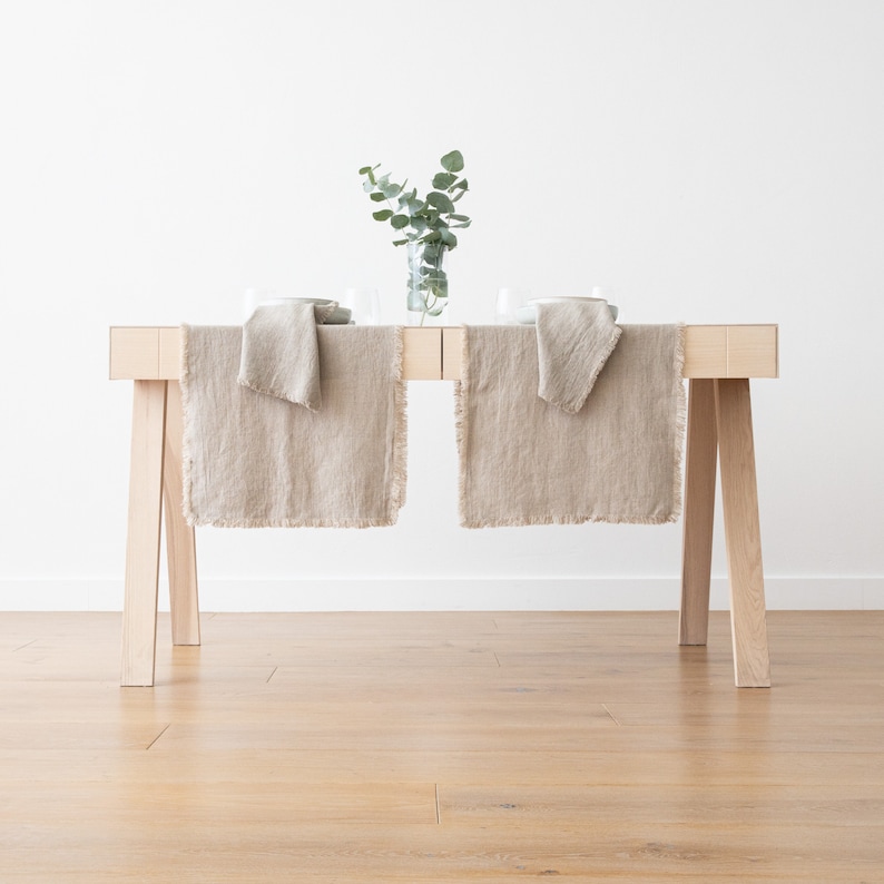 May include: A light wood table with two beige linen table runners with frayed edges. A vase with green leaves sits in the center of the table.