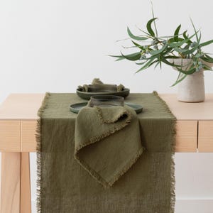 May include: Olive green linen table runner with fringed edges, set with matching plates, bowls, and a folded napkin. A vase with greenery sits on the table. The table is made of light-colored wood.