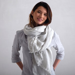 May include: A woman wearing a light gray linen scarf with frayed edges. The scarf is wrapped around her neck and tied in a loose knot. She is wearing a light gray linen shirt.