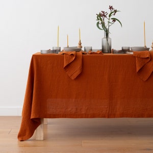 Könnte beinhalten: Ein Esstisch, gedeckt mit einer rostfarbenen Leinentischdecke. Der Tisch ist mit grauen Tellern, Servietten und Gläsern gedeckt. Hohe gelbe Kerzen und eine Vase mit Grünzeug vervollständigen die elegante Anordnung.