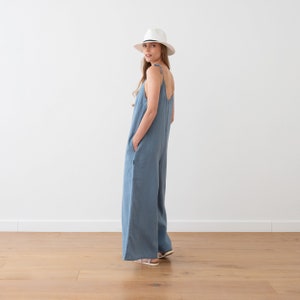 May include: A woman wearing a light blue linen jumpsuit with wide legs and thin straps. She is wearing a white fedora hat and is standing on a wooden floor.