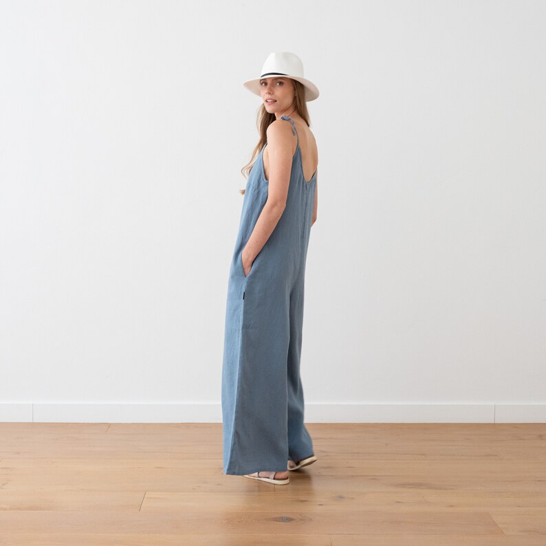 May include: A woman wearing a light blue linen jumpsuit with wide legs and thin straps. She is wearing a white fedora hat and is standing on a wooden floor.