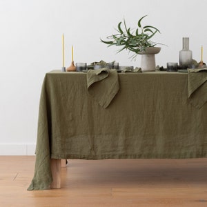 Puede incluir: Un mantel de lino verde con una servilleta doblada sobre una mesa de madera. Hay vasos y velas en la mesa, y un jarrón con hojas verdes en el centro.