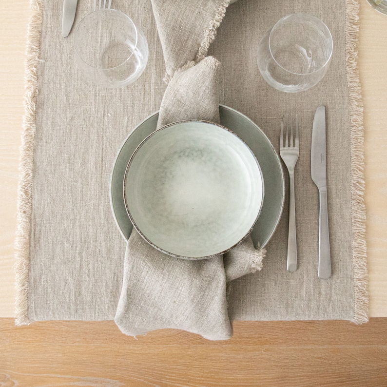 May include: A light gray ceramic bowl with a textured surface sits on a beige linen placemat with a fringe border. The bowl is surrounded by a larger, light gray ceramic plate and two silver forks and knives.