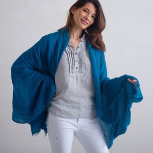 May include: A teal blue linen scarf with frayed edges. The scarf is draped over a person wearing a light gray linen shirt and white jeans.