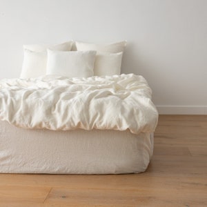 May include: A white linen duvet cover and pillowcases on a bed with a white headboard. The bedding is wrinkled and has a natural, relaxed look.