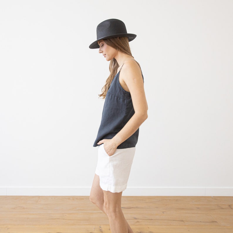 May include: A woman wearing a black fedora hat, a dark gray sleeveless top, and white linen shorts. She is standing in a room with white walls and a wooden floor.