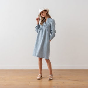 May include: A light blue linen dress with a white hat and sandals. The dress has a pleated detail at the neckline and long sleeves.