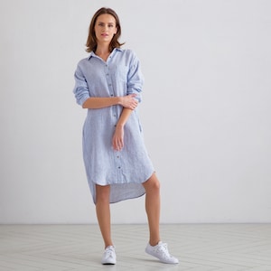 Puede incluir: Una mujer lleva un vestido camisa de lino a rayas azul claro con un dobladillo alto-bajo. Est&aacute; de pie sobre un suelo blanco y lleva zapatillas blancas.