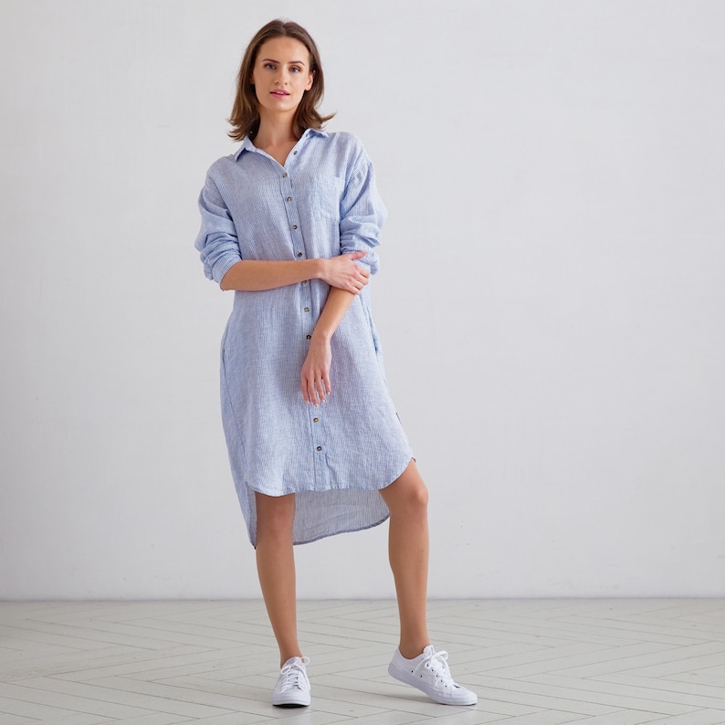 Puede incluir: Una mujer lleva un vestido camisa de lino a rayas azul claro con un dobladillo alto-bajo. Est&aacute; de pie sobre un suelo blanco y lleva zapatillas blancas.