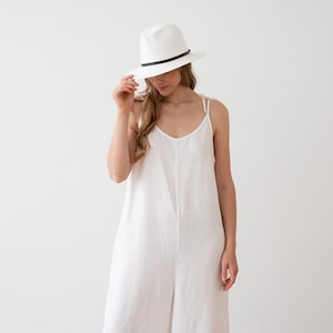May include: A white linen jumpsuit with thin straps and a relaxed fit. The model is wearing a white fedora hat with a black band.