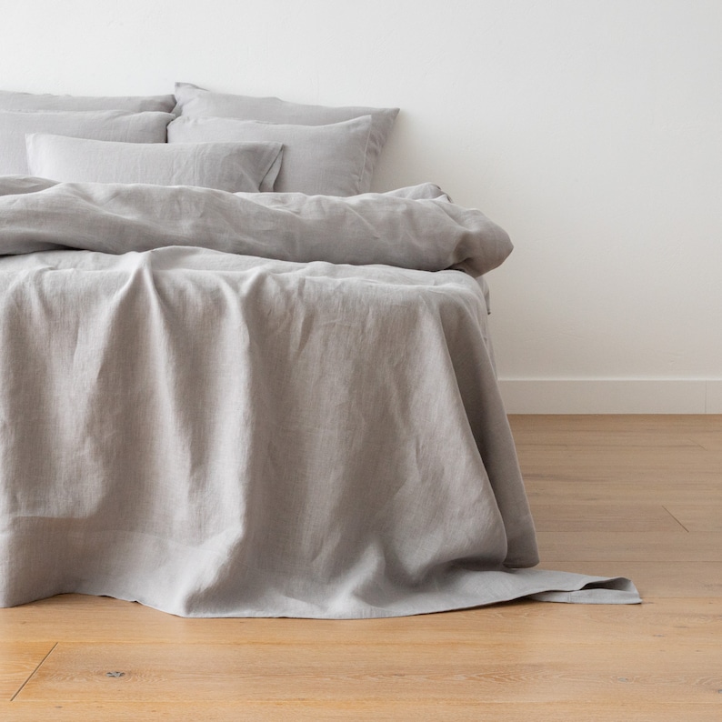 May include: A gray linen duvet cover with a fitted sheet and pillowcases. The bedding is arranged on a bed with a white headboard and a wooden floor.