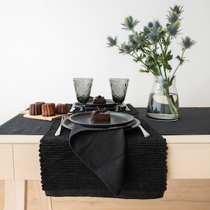 May include: A black table setting with a black placemat, black napkins, and black plates. There are two glasses of water, two small chocolate cakes, and a vase of blue thistle flowers.