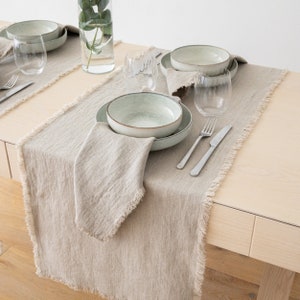 May include: A light brown linen table runner with frayed edges. The runner is laid on a wooden table and features a place setting with a light green bowl, a fork, and a knife.