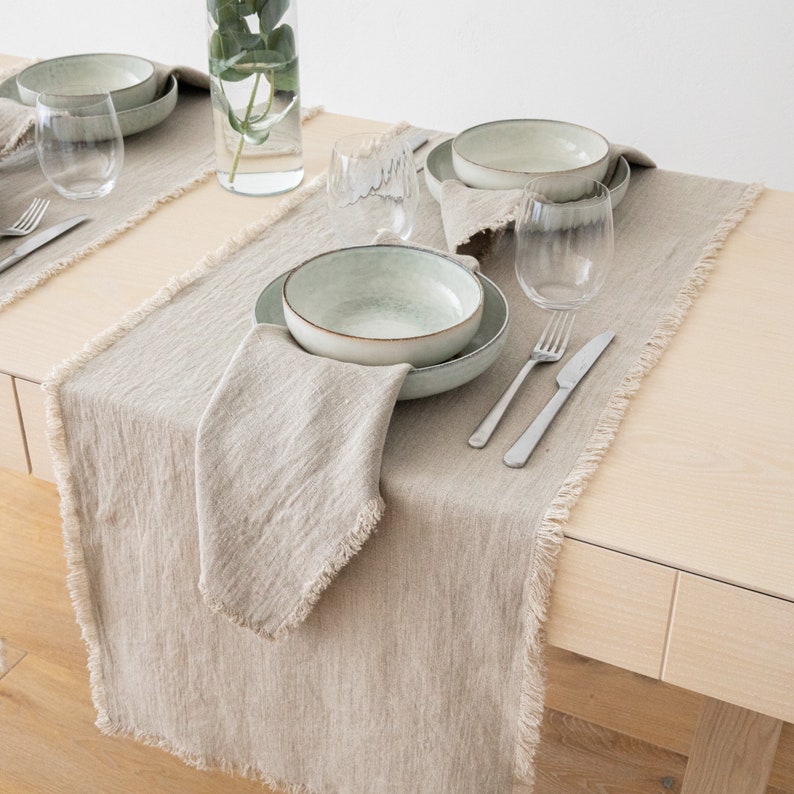May include: A light brown linen table runner with frayed edges. The runner is laid on a wooden table and features a place setting with a light green bowl, a fork, and a knife.
