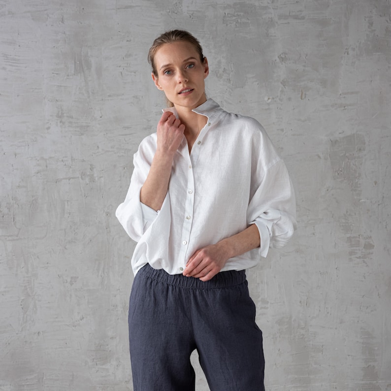 K&ouml;nnte beinhalten: Ein wei&szlig;es Leinenhemd mit Kn&ouml;pfen und langen &Auml;rmeln. Das Hemd ist leicht oversized und hat eine lockere Passform. Das Model tr&auml;gt das Hemd zu einer dunkelgrauen Leinenhose.