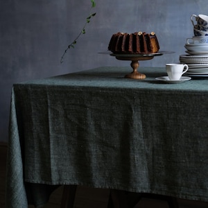 Puede incluir: Un mantel de lino verde oscuro drapeado sobre una mesa de madera con un soporte para pasteles que sostiene un pastel de chocolate con glaseado marrón. Una taza de té blanca y un platillo están sobre la mesa.