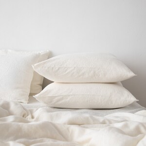 May include: Two white linen pillowcases stacked on top of a white linen bed sheet. The pillowcases are made of a soft, natural fabric and have a relaxed, casual look.