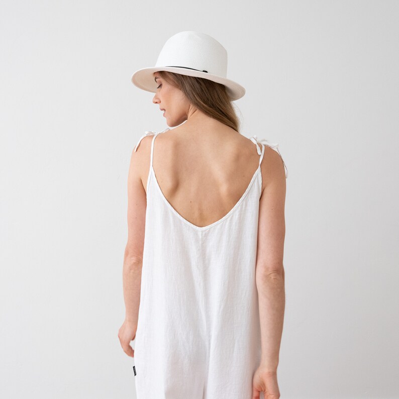 May include: A white linen jumpsuit with thin straps that tie at the shoulders. The jumpsuit has a deep V-neckline and a relaxed fit. The model is wearing a white hat.