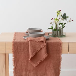 May include: A rust-coloured linen table runner with a fringed edge, alongside matching napkins, grey bowls, and plates. A clear glass vase holds a floral arrangement, creating a warm dining setting.