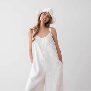 May include: A woman wearing a white linen jumpsuit with thin straps and a white wide-brimmed hat.