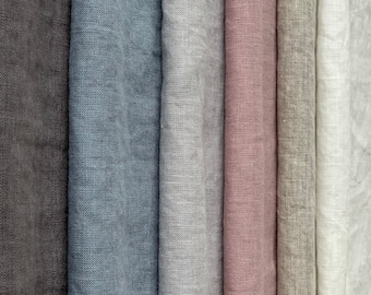 May include: A collection of vertical fabric swatches featuring a woven texture. The colours displayed from left to right include dark charcoal, slate blue, light grey, dusty rose, beige, and off-white. Each piece of fabric hangs in soft, parallel folds.