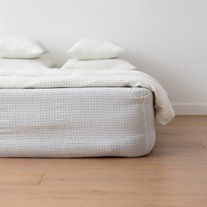 Puede incluir: Cama blanca con una sábana bajera a cuadros blanco y negro y una funda nórdica blanca. Dos almohadas blancas están encima del edredón.