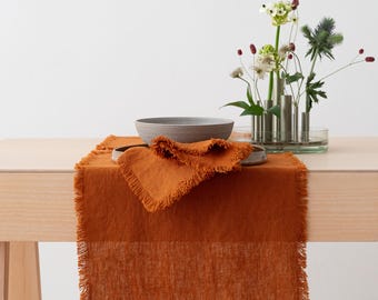 May include: An orange linen table runner with a fringed edge is displayed on a light wood table. A matching napkin and a gray ceramic bowl are placed on the runner. A vase with flowers is in the background.