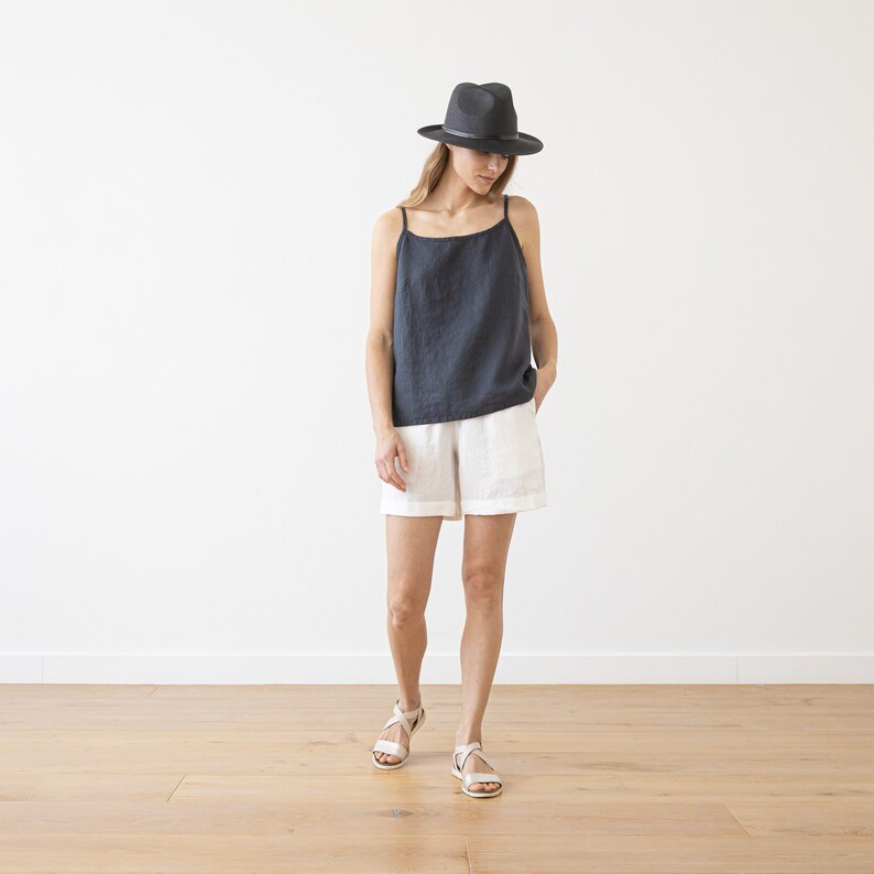 May include: A woman wearing a black linen tank top, white linen shorts, and a black fedora hat. She is standing on a wooden floor.
