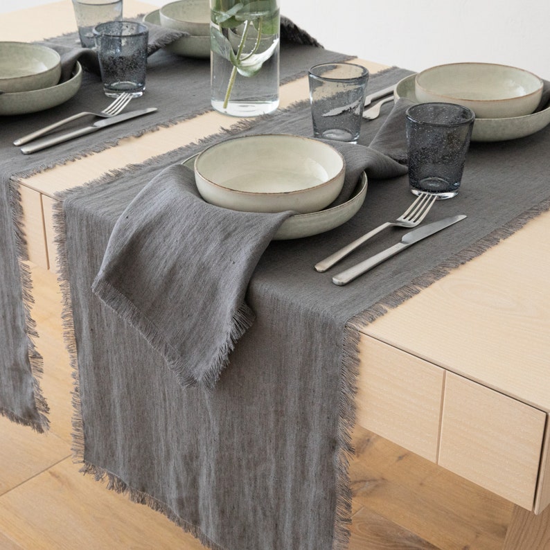 May include: A gray linen table runner with frayed edges. The runner is placed on a wooden table with a place setting of a bowl, plate, silverware, and a glass.