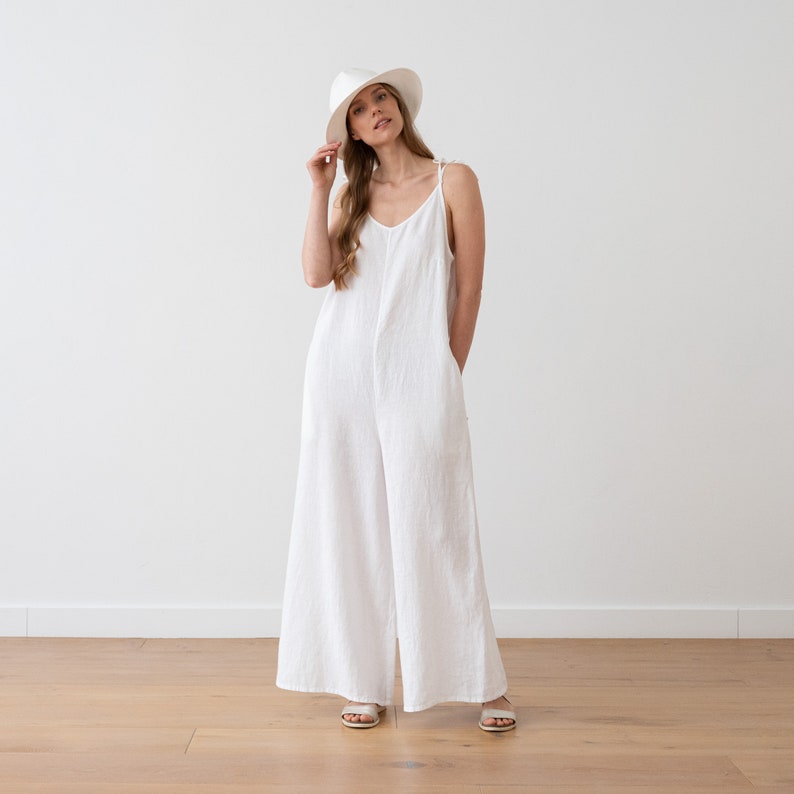 May include: A white linen jumpsuit with thin straps and wide legs. The model is wearing a white hat and sandals.